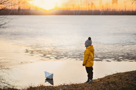 A boy in a yellow jacket is standing on the shore of a lake, a paper boat is floating nearby. Early spring, ice melts on the lake, evening, sunset, the sun sets over the horizon.