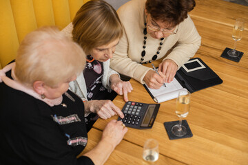 Top view of intelligent good-looking elderly women meeting in modern cafe with orange walls , having calculator and notebook on table, dicussing prices, counting and writing down figures