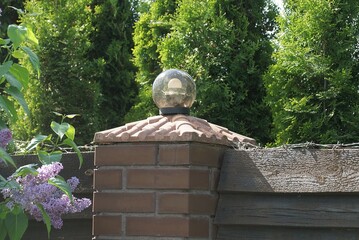 one round glass lantern with a white lamp on a brown wall of a fence made of bricks and wooden boards on the street against the background of green trees