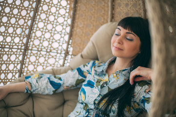 Portrait of a beautiful girl sitting in a round hanging chair. Cute brunette woman peacefully resting in armchair