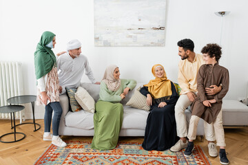 happy interracial women sitting on sofa near muslim family at home.