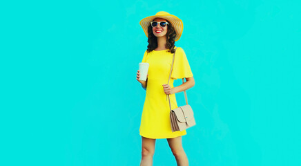 Portrait of beautiful happy smiling woman with cup of coffee wearing yellow summer hat and dress on blue background