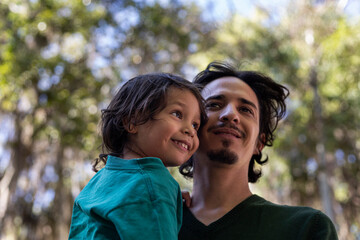 Latin American dad carries his 3 year old son in his arms in the park. Close up shot. Family and...