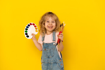 Blonde curly girl on a yellow background smiles, with paints and tassels in her hands, dressed in a...