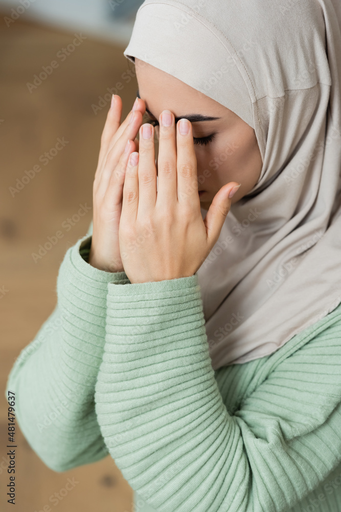 Poster young arabian woman holding hands near face while praying at home.