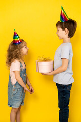 Little blonde girl accepts gift in pink box for her birthday from her brother, on a yellow background, children in caps, vertical photo