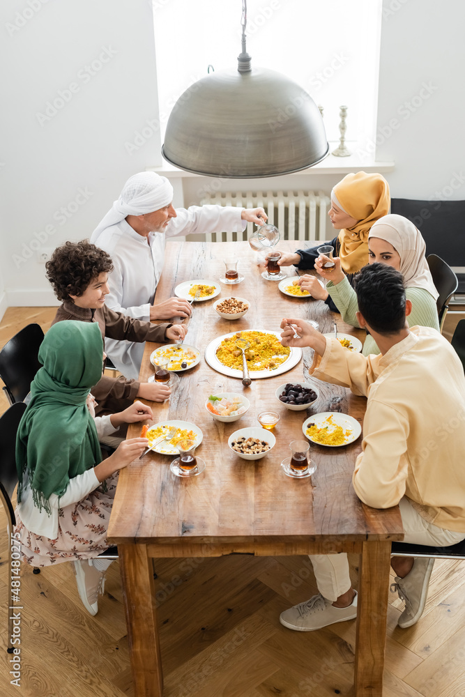 Wall mural high angle view of middle aged muslim man pouring tea during dinner with multiethnic family.