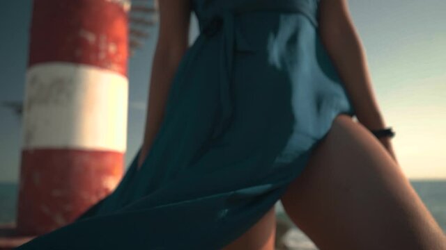 Slow motion m&eacute;dium shot, en tila de una mujer joven latina y hermosa con cabello chino casta&ntilde;o, en un vestido azul, de pie sobre las rocas en la playa, a un lado de un faro, mirando el amanecer