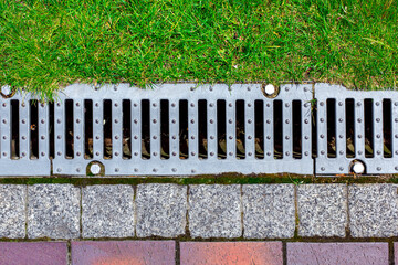 gray grating of the drainage system for drainage of rainwater in the park at the edge of the sidewalk from a stone tile with a green lawn, landscaping a city garden close-up top view, nobody. - obrazy, fototapety, plakaty