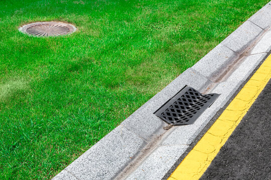 gray gutter of a stormwater drainage system on the side of asphalt road with yellow markings and sewer septic tank in green lawn, concrete drainage ditch with iron grate.
