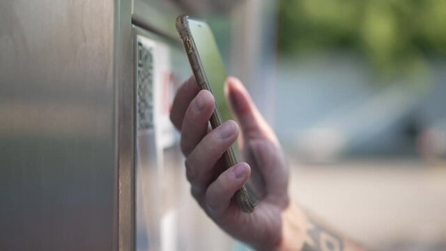 Close-up contactless payment at car wash service outdoors on sunny spring summer day. Male tattooed Caucasian hand using smartphone app paying for service. Convenience and maintenance
