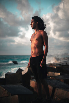 Hombre joven latino, parado en un rompeolas en la playa al amanecer, mirando al horizonte, sintiendo el viento y los rayos de sol