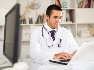 Positive man doctor sitting at workplace with computer in her office