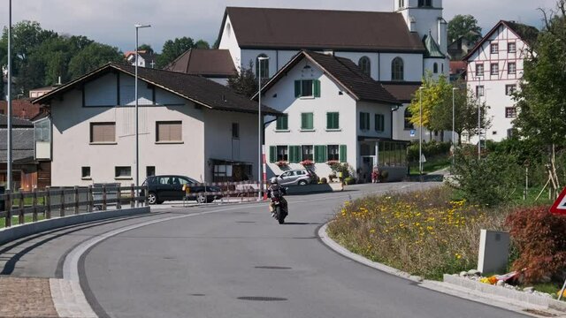Landscape asphalt road between residential Houses in Liechtenstein, Eschen. Motorcycle ride on Empty Highway by city Street in Alps Mountain Valley at Sunny Day. Biker rides by clean road in the city.