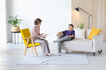 Boy sits in an imposing position on sofa, crossing his legs. The psychologist sits on opposite sides and conducts conversation with him. Full-length image.