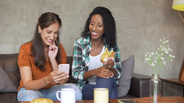 Woman scroll down mobile phone to show social media content to friend at coffee shop. Female friends laugh and have fun with cell phone sitting in couch.
