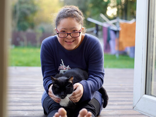 Portrait of girl with Down Syndrome sitting on patio with cat - obrazy, fototapety, plakaty