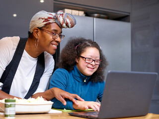 Mother with daughter with Down Syndrome looking at laptop in kitchen - obrazy, fototapety, plakaty