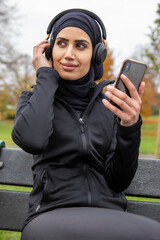 Woman in black sports clothing and hijab listening to music in park