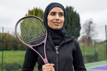 Portrait of woman in black sports clothing and hijab holding tennis racket - obrazy, fototapety, plakaty