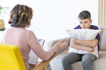 Worried teenager in purple T-shirt hugs pillow while sitting on couch with psychologist for...