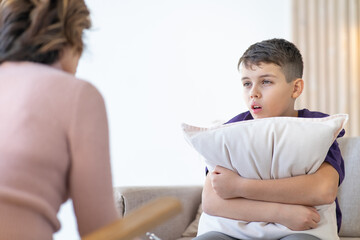 Worried teenager hugs pillow while sitting on couch with psychologist for consultation. Psychologist in chair in front of blurred background.