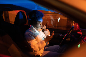 Serious black woman read bad news in smartphone sit in car at drivers seat at night. Concerned...