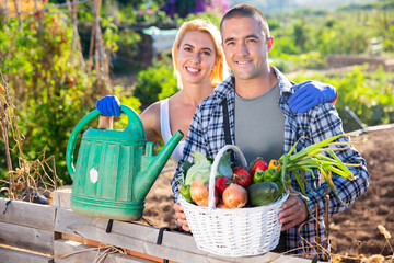 Portrait of cheerful young man and woman with basket full of freshly picked vegetables posing at garden