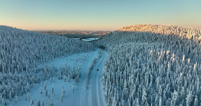 Aerial drone view following a car driving in sunlit, snow covered forest of Lapland