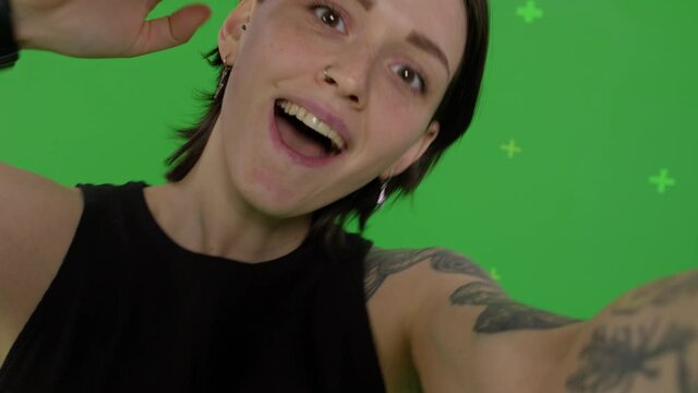 Young smiling woman posing on green screen background. Girl taking selfie self portrait photo on smartphone. Chroma key. Female model showing positive face emotions. 4k raw video footage