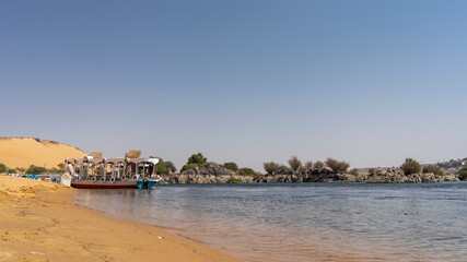 There are tourist boats on the sandy shore of the Nile. Yellow sand of the beach. Ripples on a calm transparent river. In the distance - picturesque boulders, green vegetation. Blue sky. Egypt. Aswan