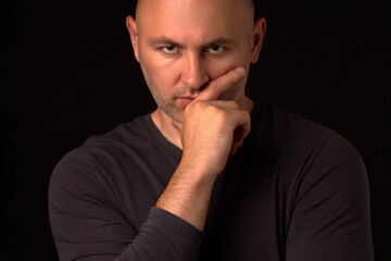 Gloomy bald man with a beard and a black sweatshirt looks thoughtfully into the camera, raising his hand to his chin. A serious man on a black background