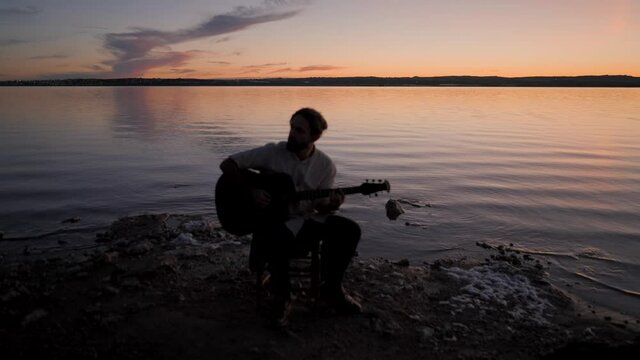 Silhouette of a guitarist playing chords on acoustic guitar by the lake on dramatic sunset in Spain  - gimbal pullback 