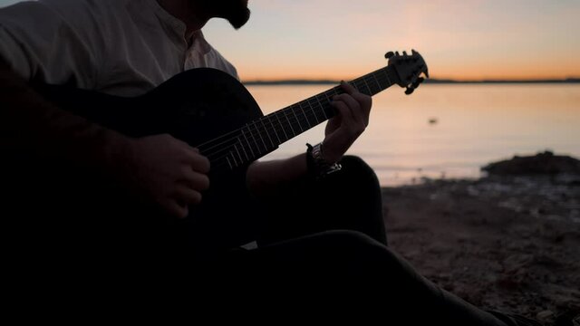 Silhouetted Hands of a man playing the guitar on the beach by the lake at golden sunset - slow-motion