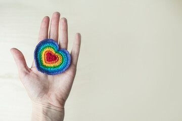 A hand in an open palm holds a knitted rainbow heart on a light neutral background