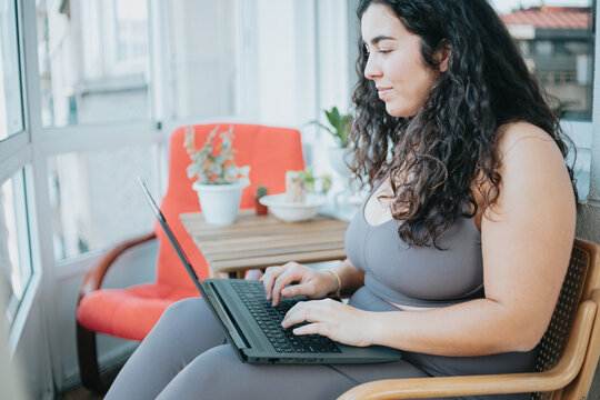 Young plus size woman in sportswear searching a using a laptop while sitting on a trendy modern flat. Preparing his diet to losing weight gaining health at home. New habit attitude