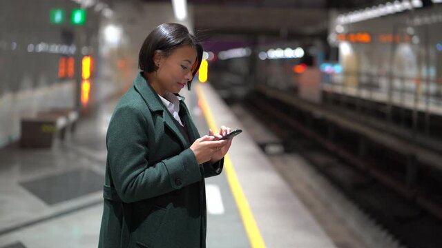 Young smiling african american woman at subway platform with smartphone scrolling social media while waiting for train, surfing web or chatting online. Digital addiction. Transportation concept