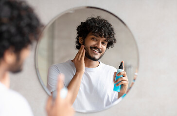 Handsome indian man applying moisturizing lotion and touching beard, smoothing after shaving in...