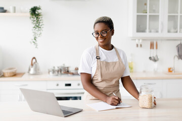 Cheerful millennial pretty black female in glasses, apron writes recipe looks pc in minimalist kitchen interior