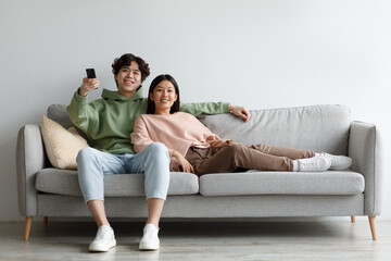 Fototapeta na wymiar Young Asian couple relaxing on sofa, watching TV, switching channels with remote control, enjoying movie at home