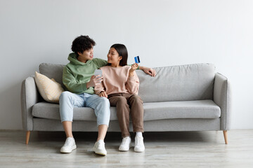 Obraz premium Young married Asian couple with credit card and smartphone sitting on couch, buying in internet store at home