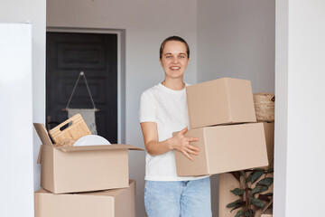 Indoor shot of happy smiling woman wearing white casual style T-shirt and jeans standing with cardboard boxes with belongings, looking at camera with positive expression, being glad to relocate.
