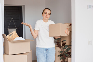 Portrait of puzzled woman wearing white casual style T-shirt and jeans standing with cardboard box...