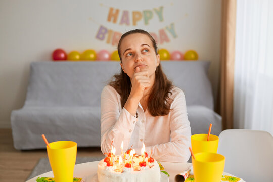 Portrait Of Sad Lonely Young Adult Thoughtful Woman Sitting Alone At Birthday Table With Cake, Holding Chin And Looking Away, Having Pensive Expressing, Thinking About Birthday Wish.