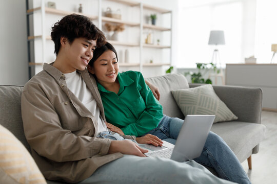Weekend pastimes concept. Married young Asian couple using laptop at home, relaxing on couch together, copy space