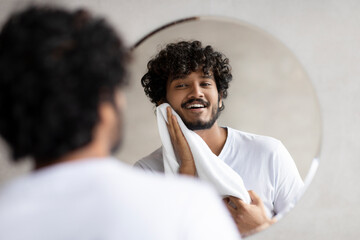 Everyday routine concept. Excited indian man wiping his face with towel while standing in bathroom,...