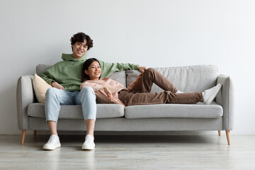 Cheerful millennial Asian couple cuddling on couch, spending weekend together at home, free space