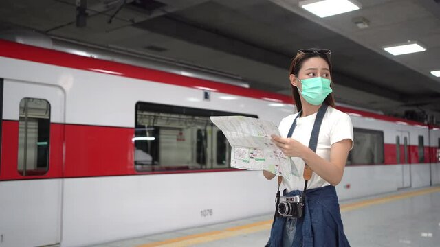 Selective focus of lost female Asian tourist with camera, wearing face mask to protect Covid-19 or Omicron, holding a map and looking around to find directions with a worried face at a subway platform