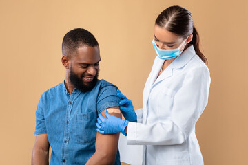 Young doctor in face mask putting band aid on black male patient's arm after covid vaccine shot on...