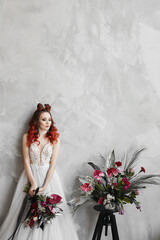 Fashionable portrait of a model girl with red hair and trendy makeup in a wedding dress posing with bridal bouquet